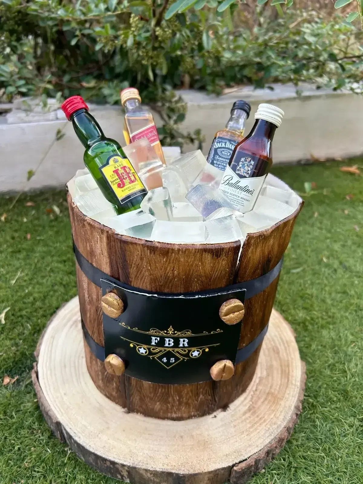 Cubo de hielo de madera con botellitas de licor surtidas sobre un soporte de tronco al aire libre.