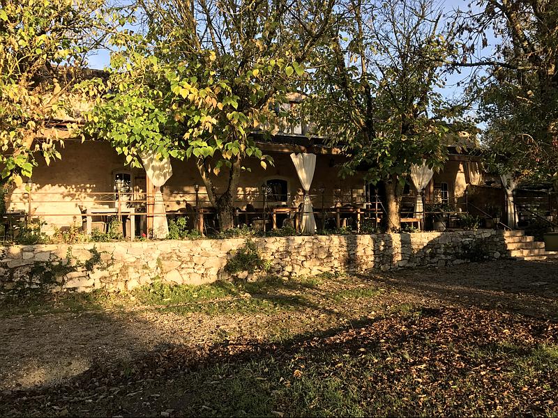 Un bâtiment rustique avec un espace repas extérieur sous les arbres, des murs en pierre et une pelouse à l'avant.