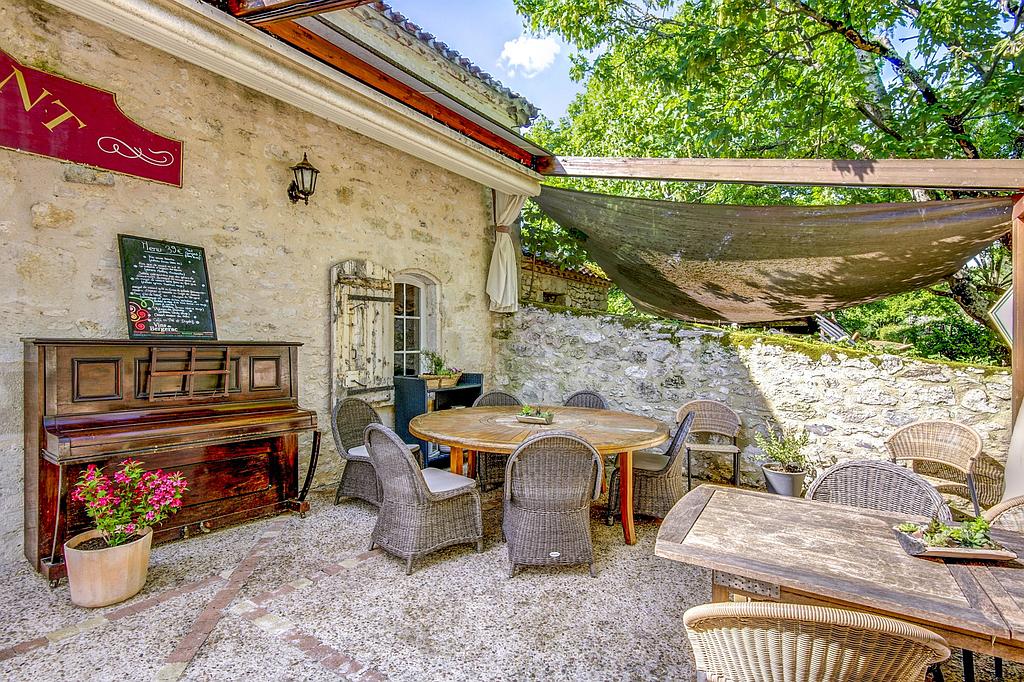 Espace repas extérieur avec tables, chaises en osier et piano. Murs en pierre, auvent et verdure.