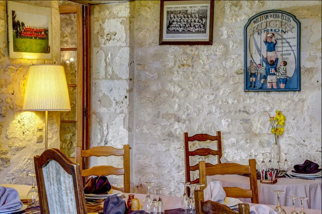 Intérieur d'un restaurant avec murs en pierre, chaises en bois, table dressée et œuvres d'art encadrées.