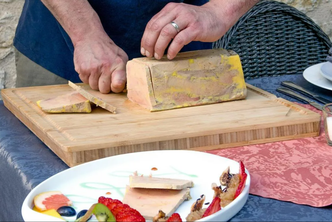 Une personne découpe du foie gras sur une planche en bois ; les tranches sont présentées avec des fruits devant.