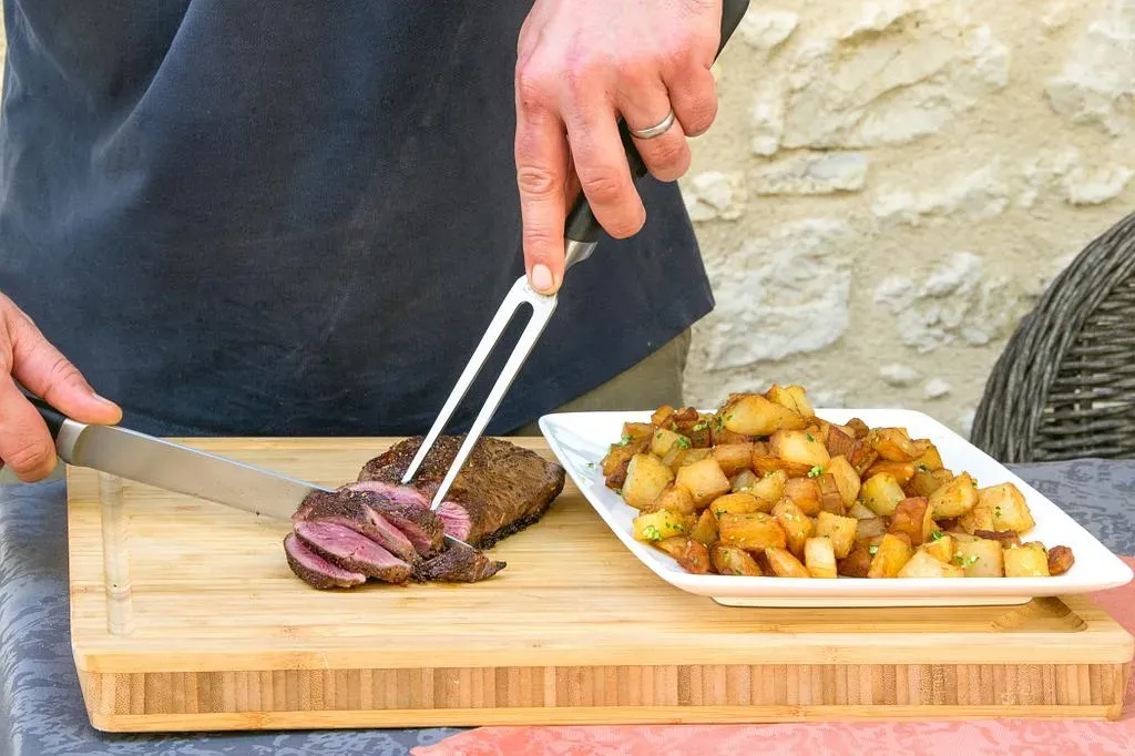 Une personne découpe de la viande cuite avec des pommes de terre sur une planche en bois à l'extérieur.