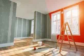 Habitación vacía recién pintada con una escalera de mano naranja, un cubo de pintura y luz solar que entra por grandes ventanas.