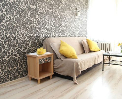 Luminoso salón con sofá beige, cojines amarillos, papel pintado estampado y mesa auxiliar junto a una ventana soleada.