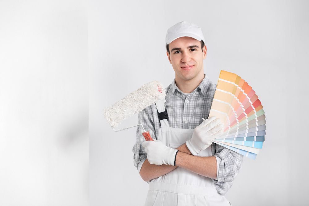 Pintor sosteniendo muestras de color y un rodillo de pintura, vestido con ropa de trabajo blanca sobre un fondo blanco.