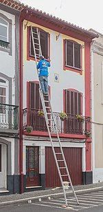 Un trabajador subido a una escalera limpia una ventana del segundo piso de un edificio rojo y blanco.