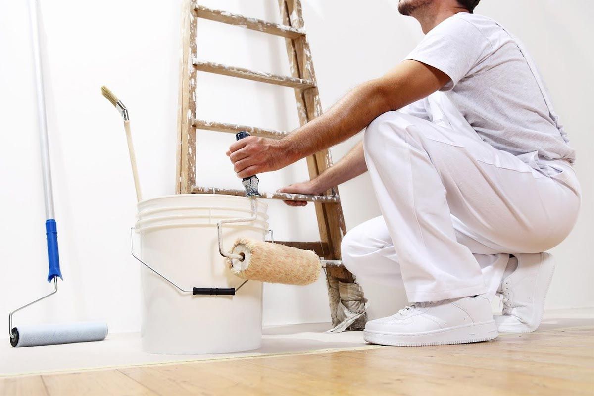 Un pintor arrodillado junto a un cubo blanco, aplicando pintura blanca fresca en una pared con una escalera al fondo.