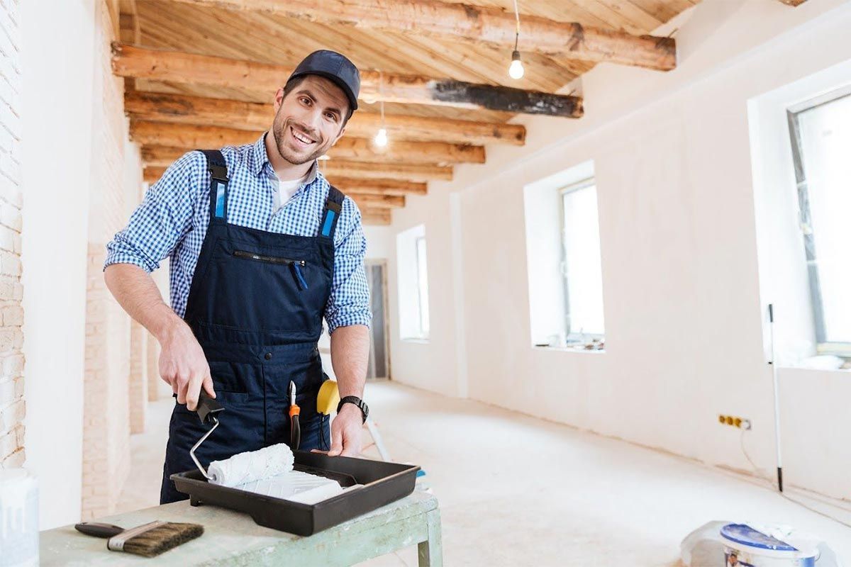 Un pintor sonríe mientras extiende la pintura en una habitación luminosa y sin terminar, con vigas de madera a la vista.