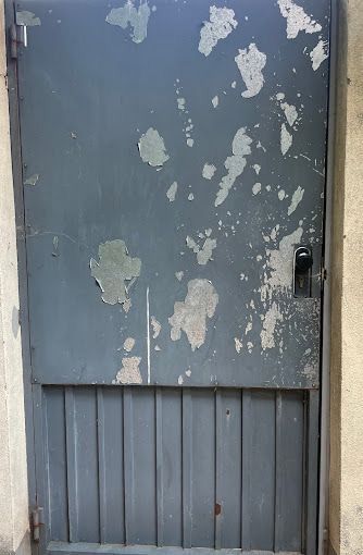 Puerta de metal azul desgastada con pintura descascarada y una pequeña manija, colocada en una pared beige.