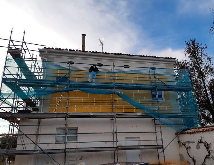 Trabajadores en andamios envolviendo una casa con una red de seguridad azul durante una renovación exterior.
