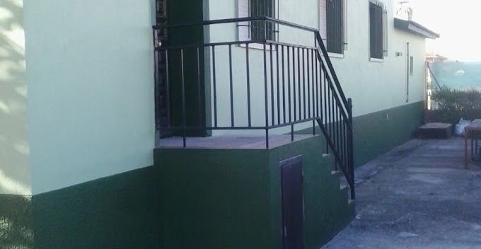 Lateral de un edificio de color verde pálido con un pequeño porche, barandillas negras y un camino de entrada inclinado que conduce a su lado.