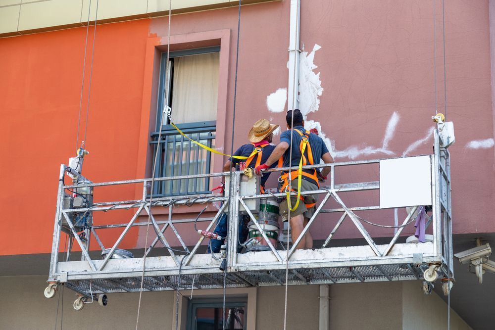 Trabajadores en un andamio suspendido reparan la fachada rosa de un edificio cerca de una ventana.