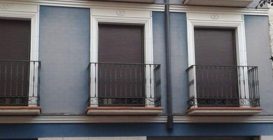 Tres ventanas cerradas con contraventanas oscuras y estrechos balcones negros en la fachada de un edificio de color azul grisáceo.