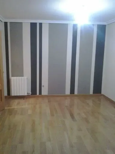 Habitación vacía con papel pintado a rayas grises, suelo de madera, radiador blanco y luz de techo.