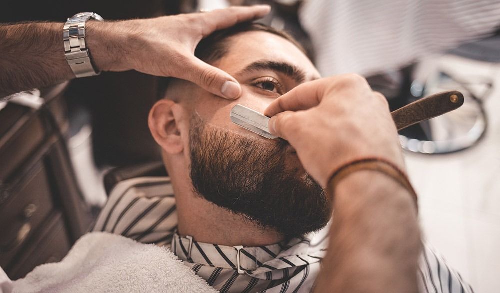 Un hombre se está afeitando la barba con una navaja por un barbero.