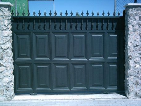 Puerta de entrada de metal gris oscuro con paneles decorativos y remate puntiagudo, situada entre columnas de piedra.