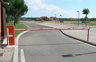 Puerta de seguridad roja y blanca que da a una calle asfaltada; casas al fondo. Día soleado.