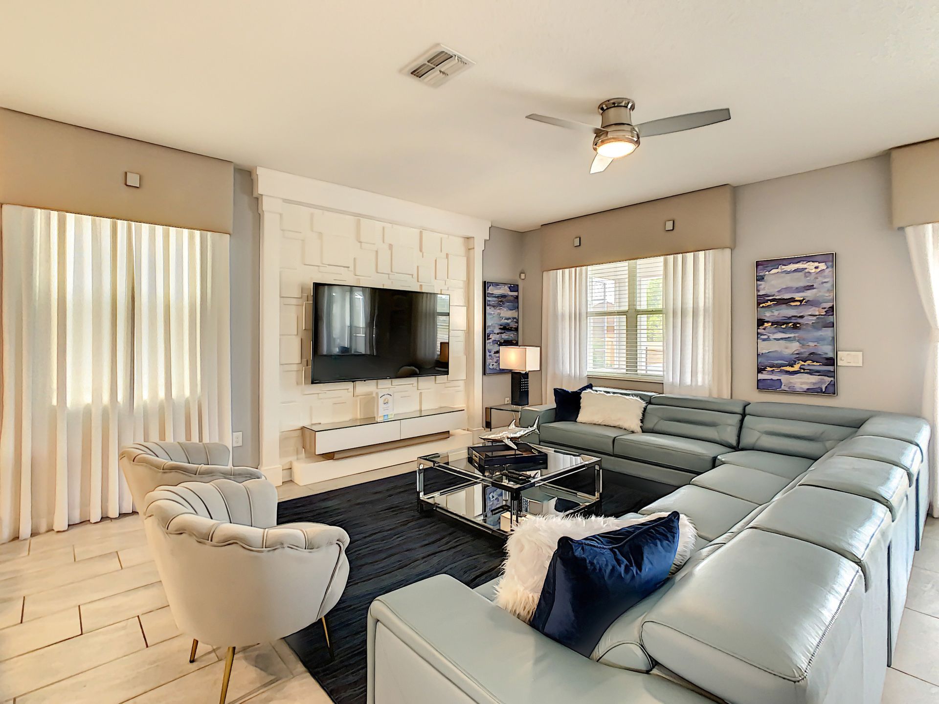 A living room with a large sectional couch , chairs , a flat screen tv and a ceiling fan.