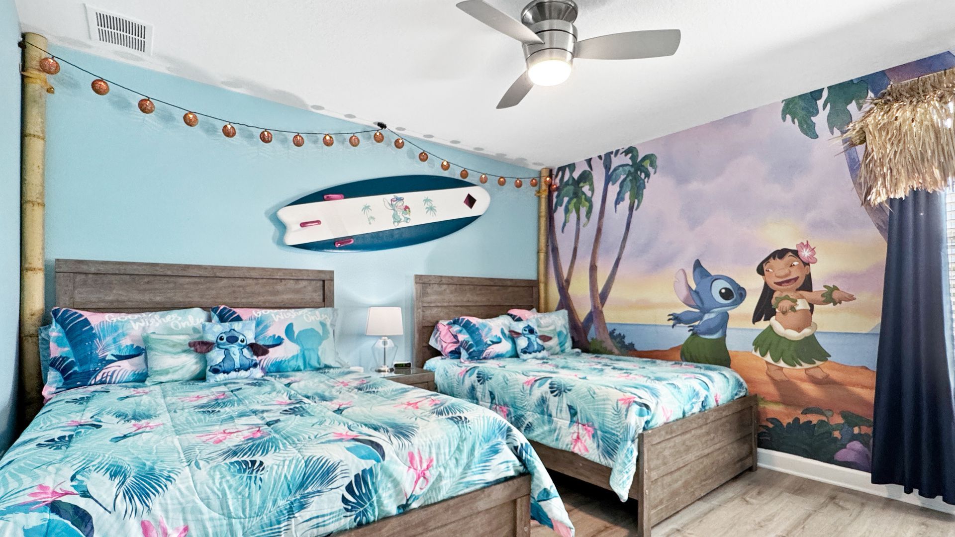 A lilo stitch bedroom with two beds , a ceiling fan and a painting of lilo and stitch on the wall.