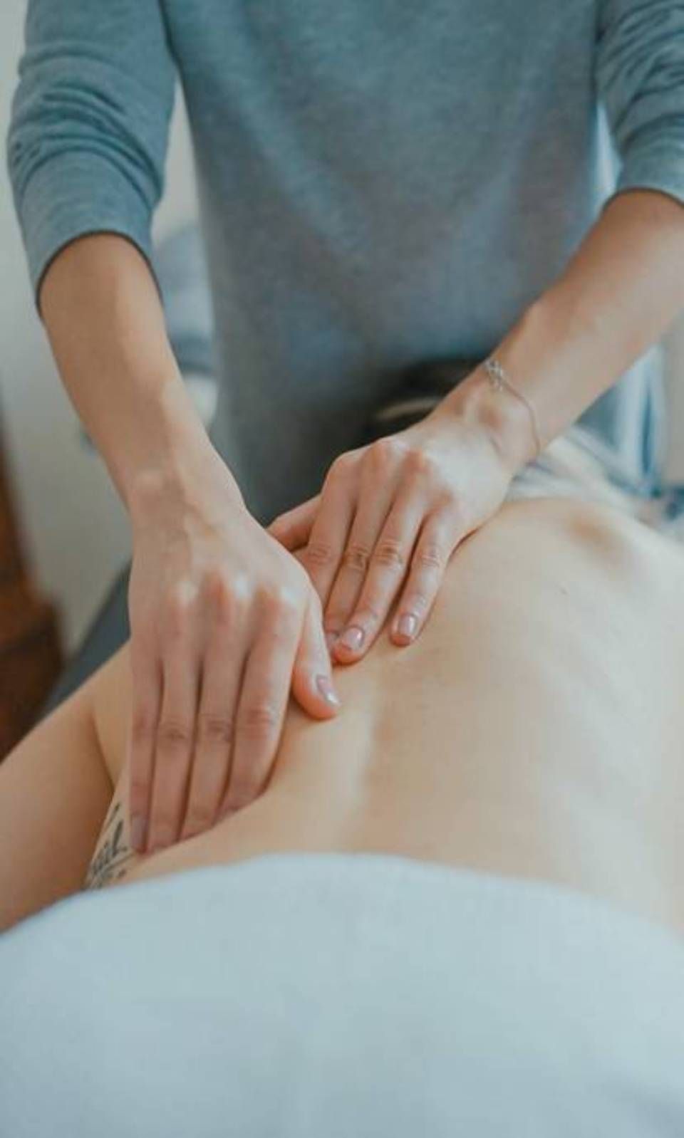 Hände massieren den oberen Rücken einer Person. Blaugraues Hemd, helle Haut, Nahaufnahme auf einer Massageliege.