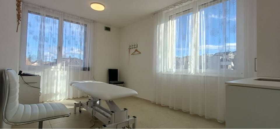 Ein heller, weisser Untersuchungsraum mit grossen Fenstern, Vorhängen und einer Massageliege.