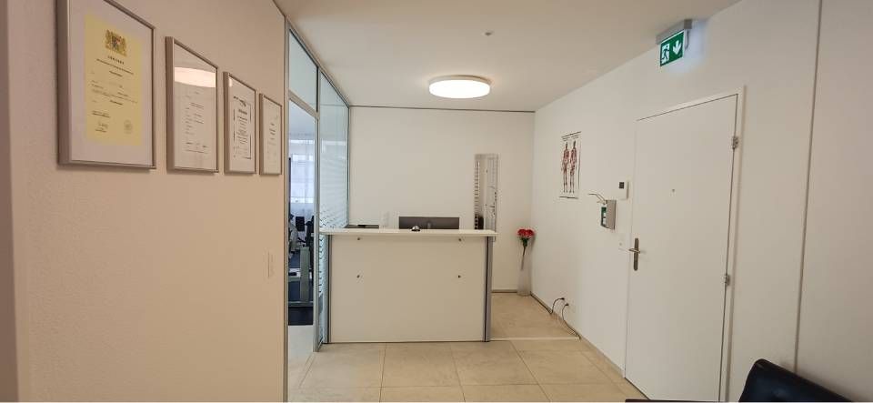 Heller Empfangsbereich mit weissem Schreibtisch, gerahmten Dokumenten, Glastrennwand und Tür mit Notausgangsschild.