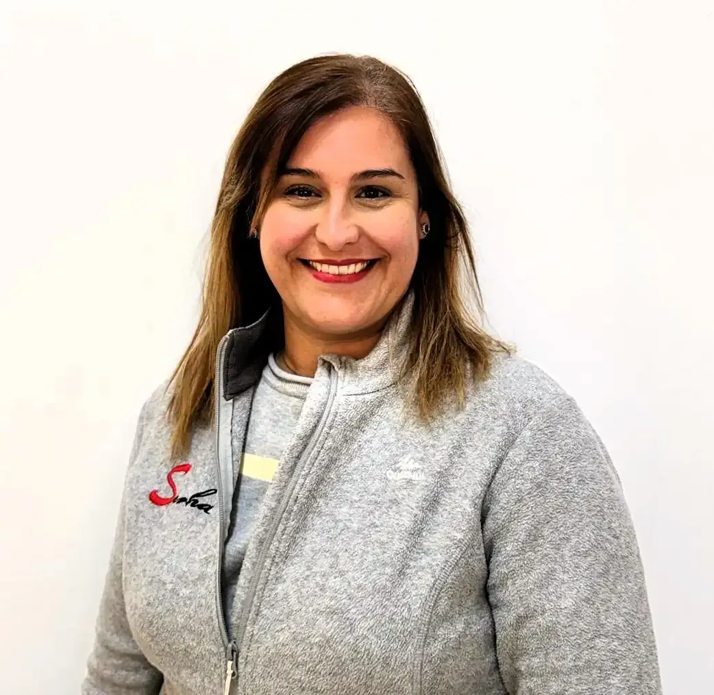 Una mujer con una chaqueta de lana gris está parada frente a una pared blanca, con el pelo ondulado.