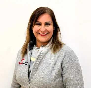 Una mujer con una chaqueta de lana gris está parada frente a una pared blanca, con el pelo ondulado.
