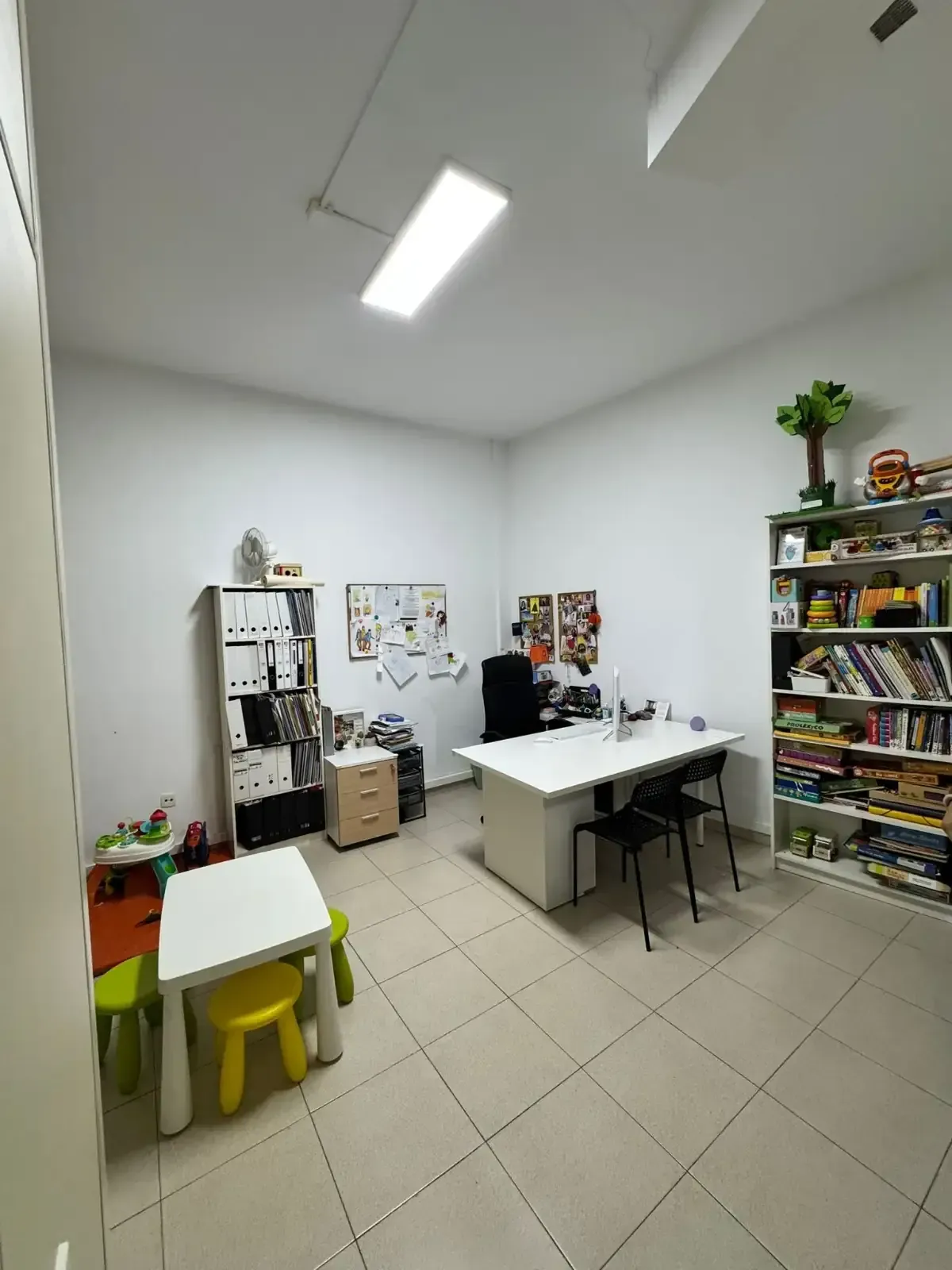 Oficina con paredes blancas, escritorio, estantes con libros, mesa pequeña y sillas.