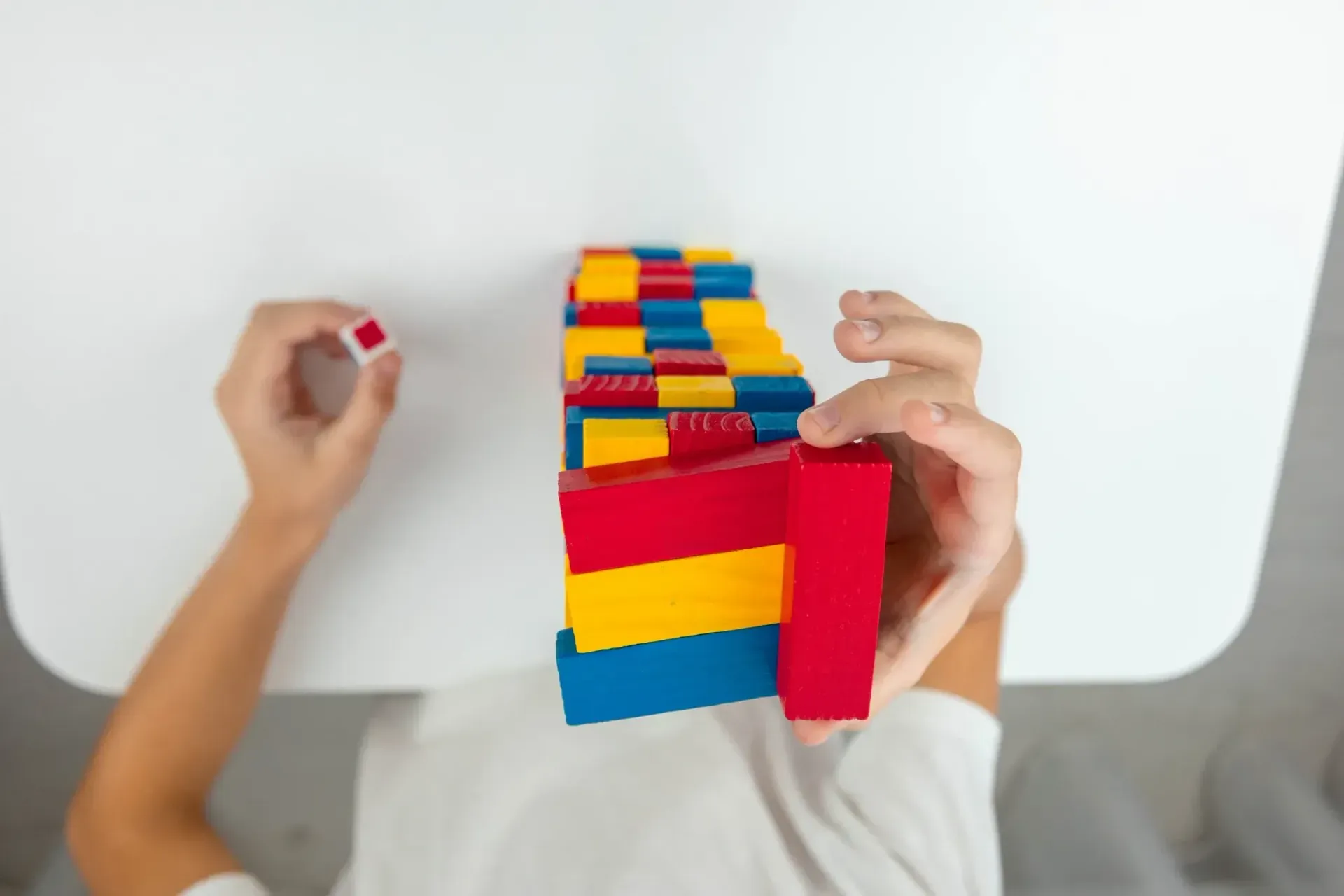 Manos jugando Jenga con bloques de madera de colores sobre una mesa blanca.