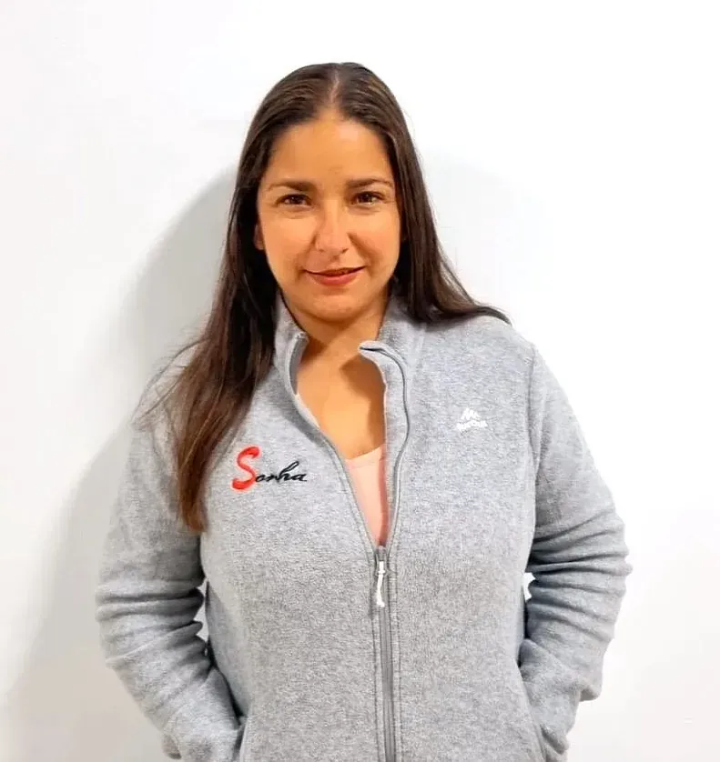 Una mujer con una chaqueta gris con cremallera sonríe a la cámara, de pie frente a un fondo blanco.