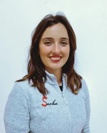 Una mujer de cabello oscuro sonríe, vestida con una sudadera gris con cremallera y la palabra 