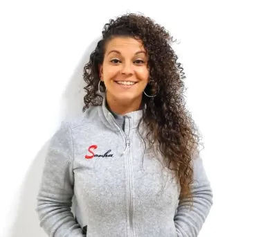 Una mujer con cabello rizado sonríe, vistiendo una chaqueta gris con cremallera y logotipo sobre fondo blanco.