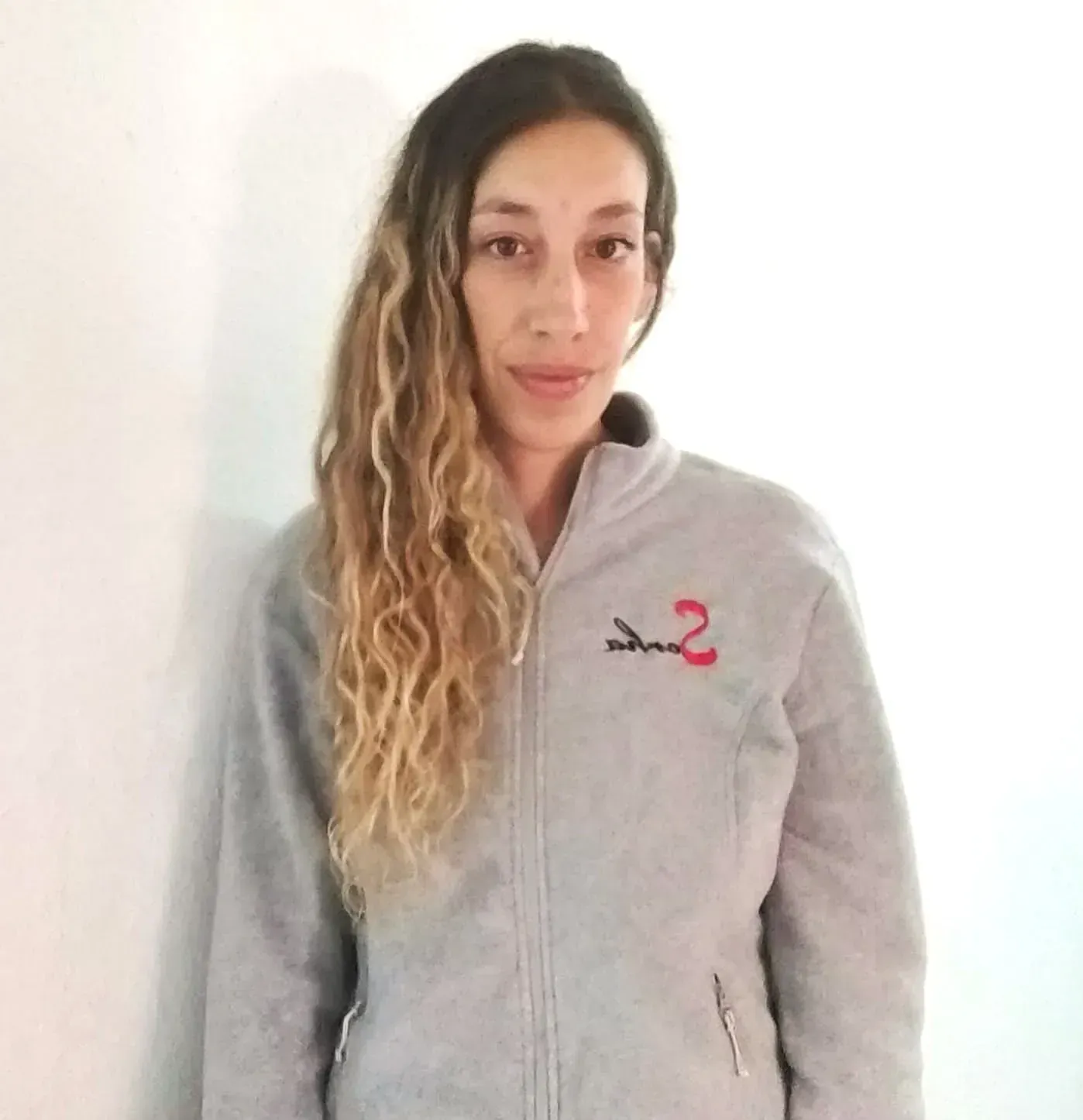 Una mujer con una chaqueta de lana gris está parada frente a una pared blanca, con el pelo ondulado.