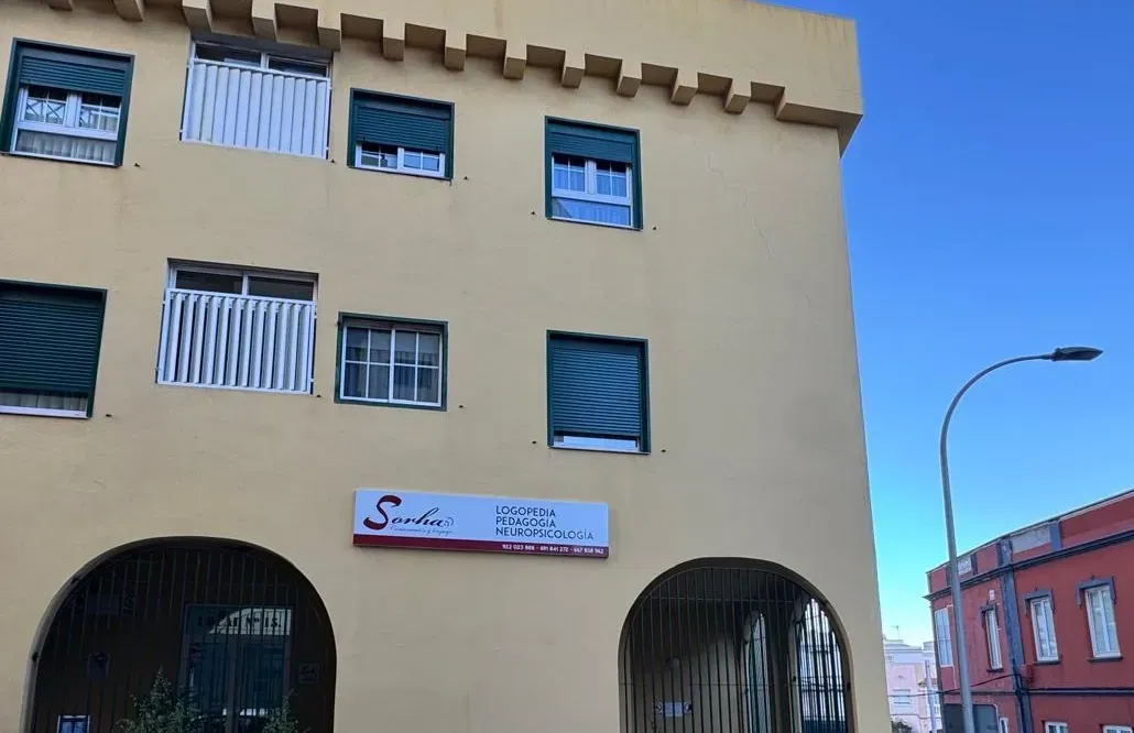 Edificio amarillo con entradas arqueadas, ventanas con persianas y un cartel que dice 