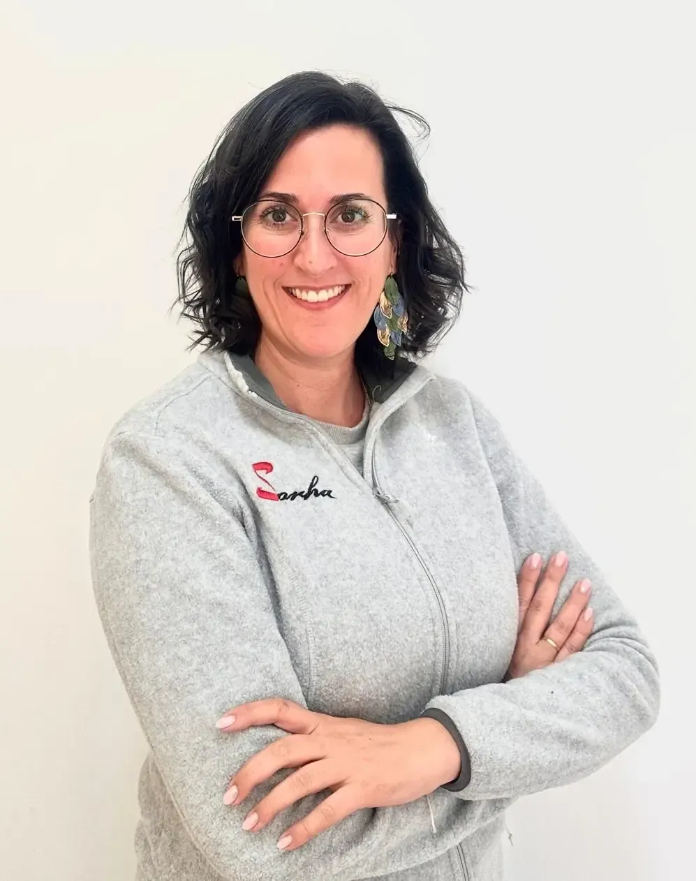 Una mujer con una chaqueta gris con cremallera sonríe a la cámara, de pie frente a un fondo blanco.