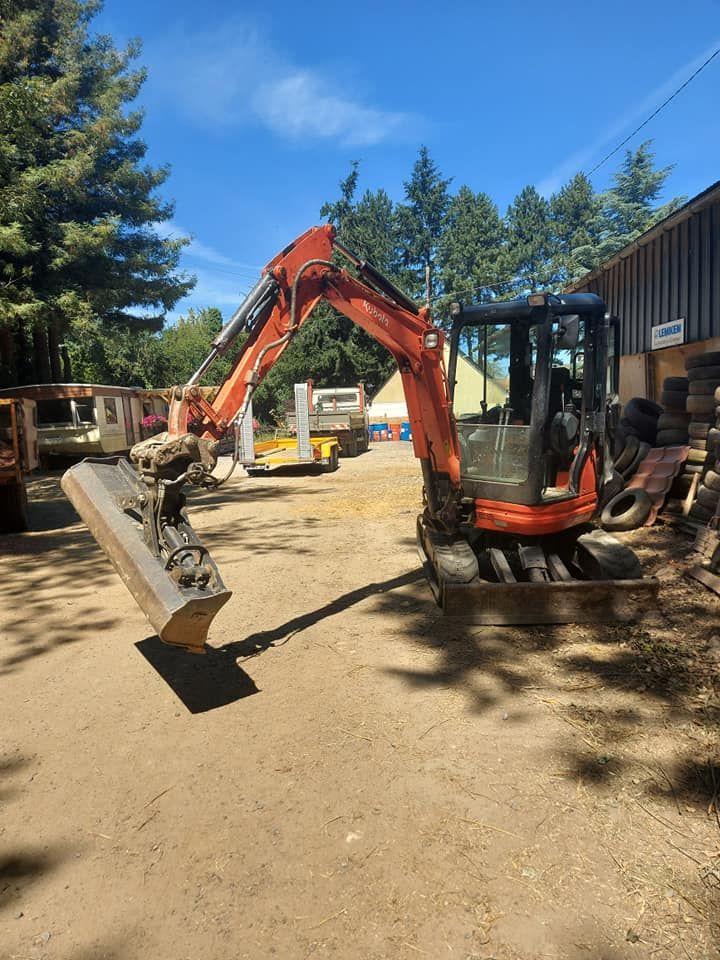 Mini-pelle orange sur un terrain meuble, devant un bâtiment et des arbres.