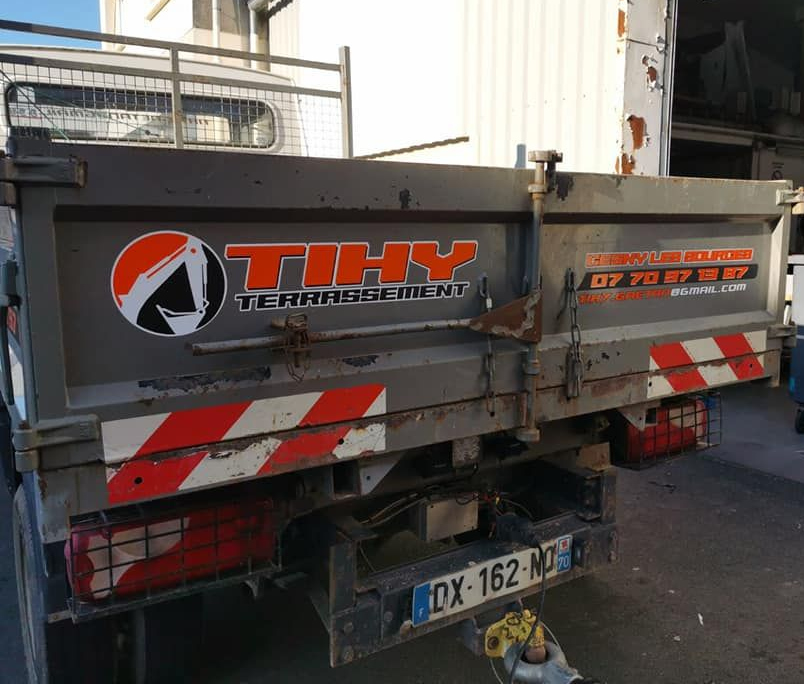 Vue arrière d'un camion-benne gris avec logo de l'entreprise et bandes de sécurité orange et blanches.