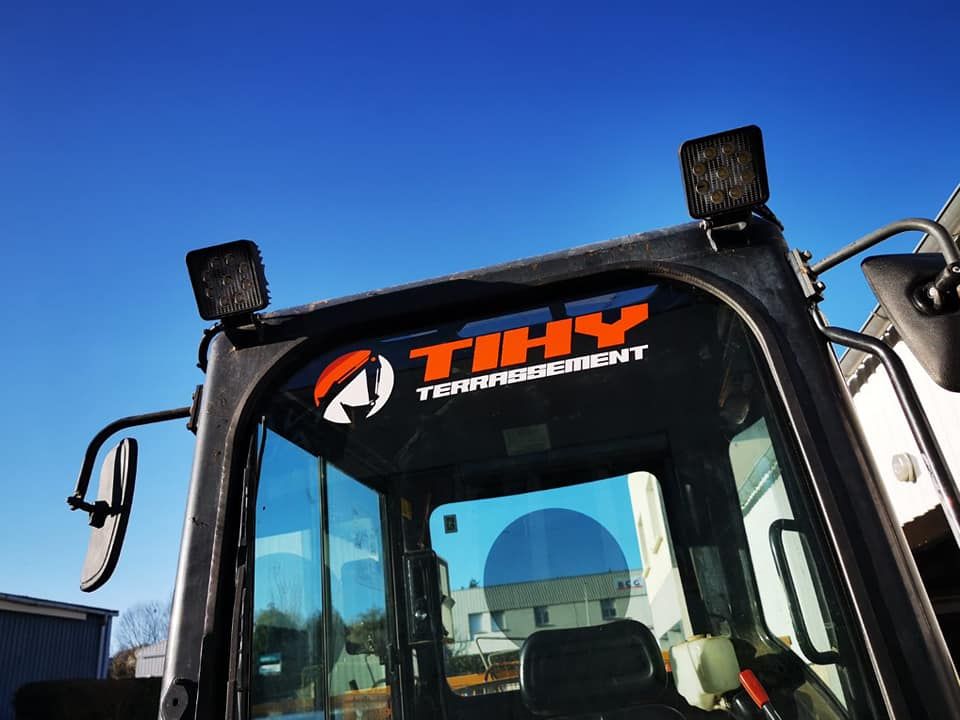 Cabine d'un véhicule de chantier noir avec le logo TIHY TERRASSEMENT et des gyrophares allumés.