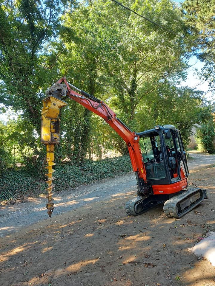 Une excavatrice orange et noire avec une tarière, sur un chemin de terre, à côté d'arbres.