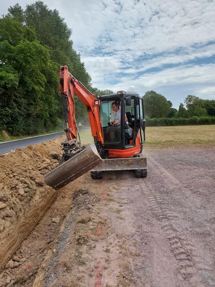Une pelleteuse orange creuse une tranchée, le conducteur est visible, le long d'une route goudronnée et d'une zone herbeuse.