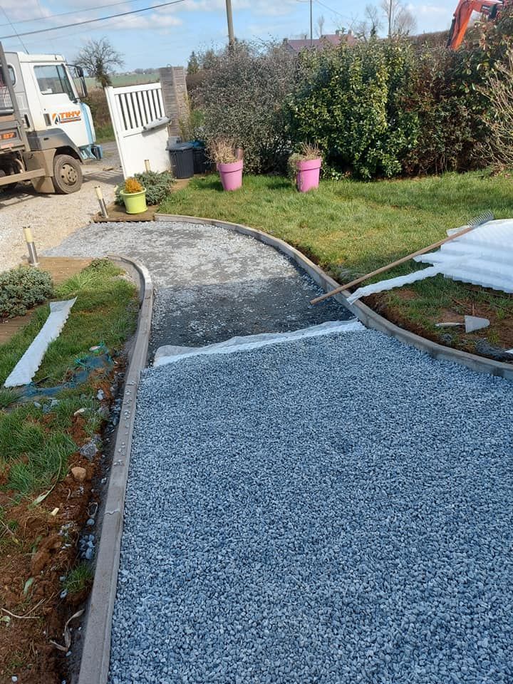 Allée de gravier en construction, bordée de bois, avec pelouse et aménagements paysagers en arrière-plan.