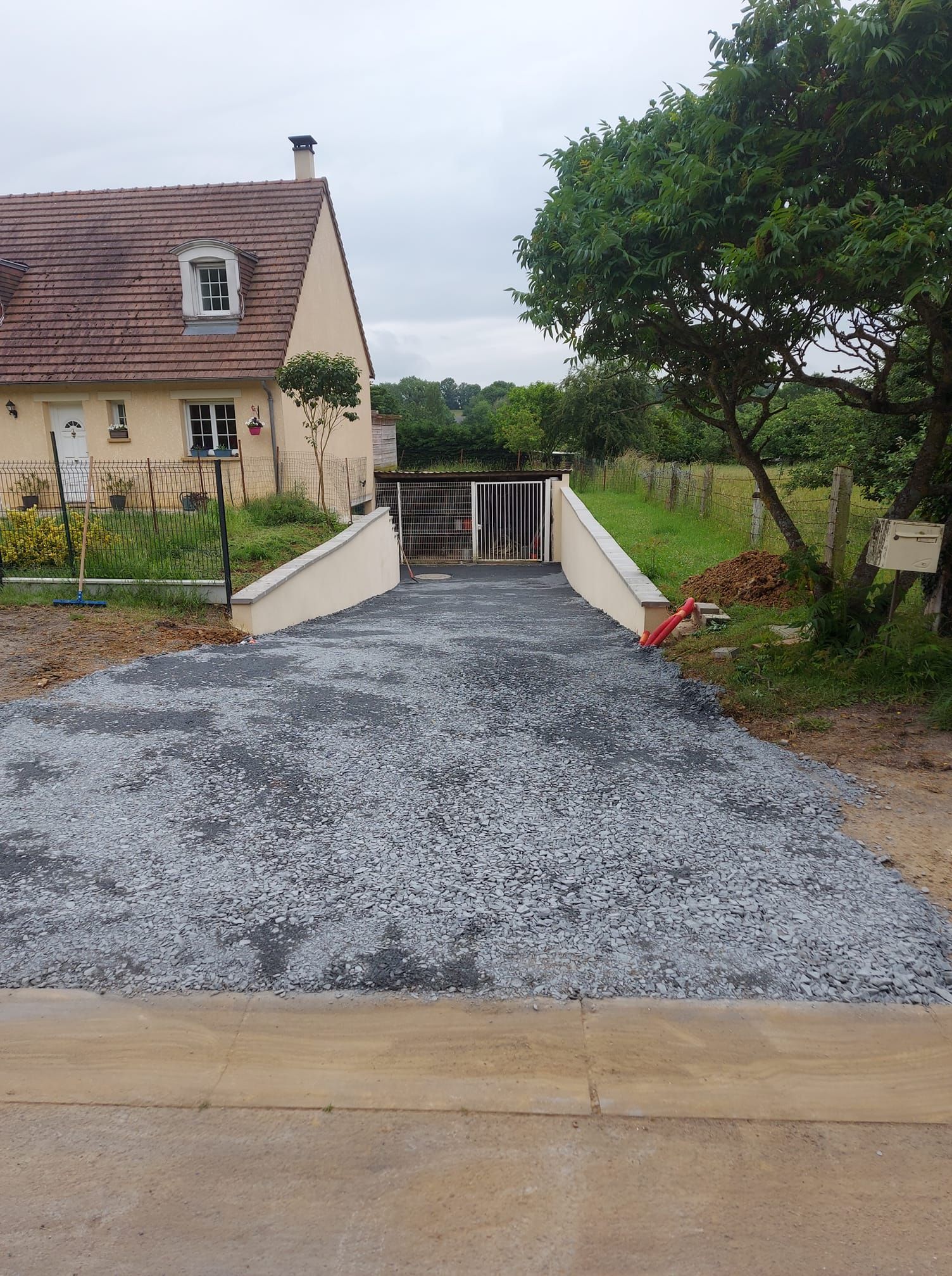 Allée de gravier menant à un portail et à une maison. Des murs couleur crème bordent l'allée.