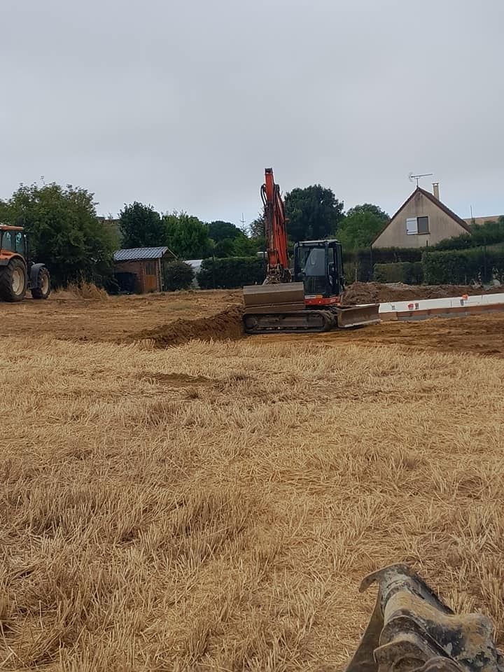 Une excavatrice creuse dans un champ, un tracteur est visible en arrière-plan.