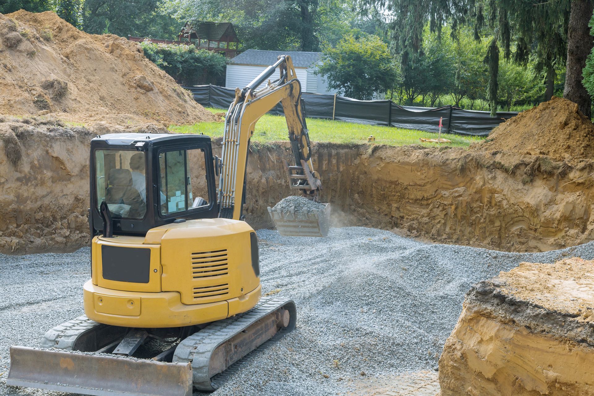 Une pelleteuse jaune creuse sur un chantier de construction avec du gravier, des tas de terre et de l'herbe verte.