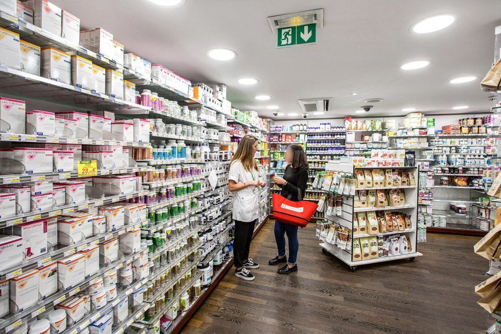 Une allée de la pharmacie avec des étagères remplies de marchandises et deux clients debout sous les lumières vives du magasin.