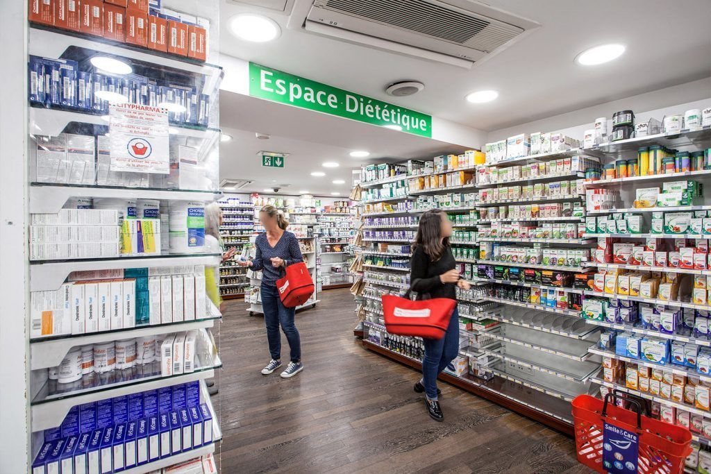 Allée de pharmacie avec des étagères de médicaments et deux clients poussant des chariots sous une lumière fluorescente vive