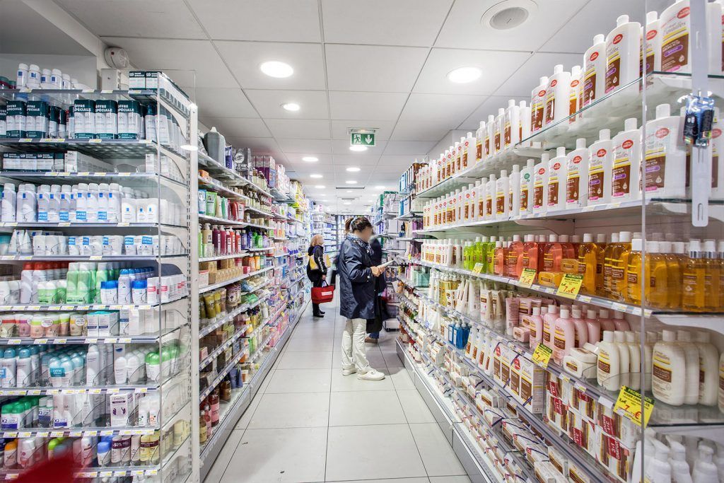 Allée lumineuse de la pharmacie avec des étagères remplies d'articles de toilette et de produits d'entretien, et quelques clients au centre.