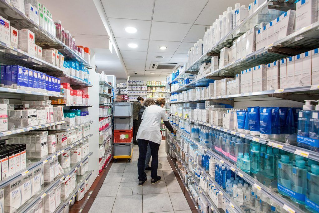Une personne en blouse blanche range des produits dans une allée de pharmacie bien éclairée, bordée d'étagères de médicaments des deux côtés.