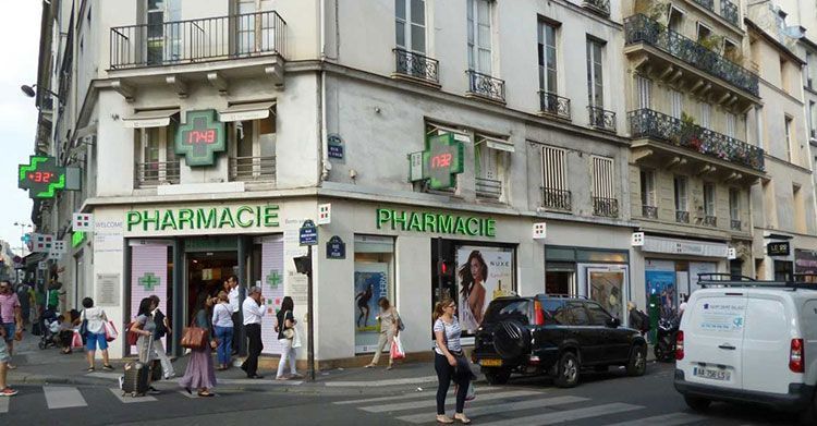Pharmacie d'angle en centre-ville, avec des passants et des voitures garées devant.